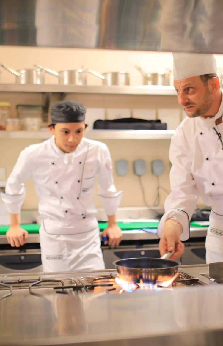 A station chef is training two apprentices at a cooking station in a restaurant kitchen.