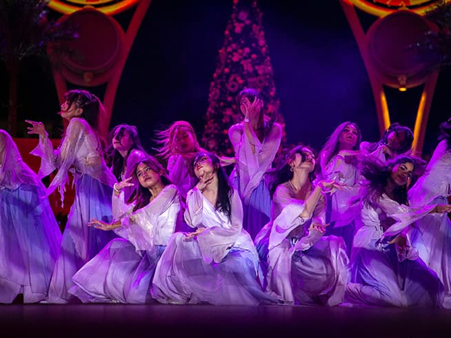 A group of dancers in white costumes perform on a stage under purple lights